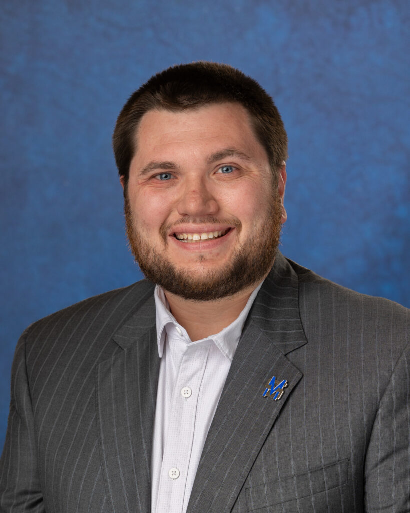 A photograph of Tracey Hickman in a light-colored shirt and gray pinstripe jacket against a blue background.